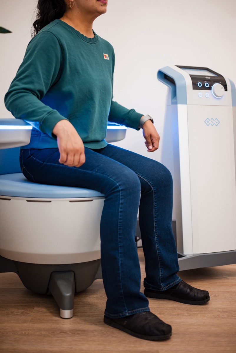 A patient seated fully clothed in the Emsella chair beside its console